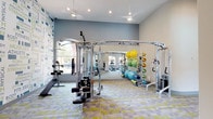 This single-floor fitness center is thoughtfully designed to support a full range of workout routines. The space features high ceilings with recessed lighting and ceiling fans, ensuring excellent airflow and visibility throughout. A large wall-to-wall mirror expands the sense of space and allows users to monitor their form during exercise. The flooring consists of a colorful geometric patterned tile, adding a dynamic and energetic aesthetic to the environment. One wall is adorned with an eye-catching typographic mural featuring fitness-related words, reinforcing an athletic atmosphere. Natural light filters in through windows, brightening the space and creating an inviting workout environment. The facility is equipped with a multi-functional cable machine and weight station at its center, complemented by a dedicated stretching and accessory area stocked with stability balls, foam rollers, resistance bands, and yoga mats in vibrant colors. Additional cardio equipment is visible toward the rear of the space. The overall design strikes a balance between functionality and modern aesthetics, making it a well-rounded destination for fitness enthusiasts of all levels.