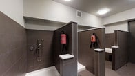 This purpose-built dog wash facility offers a clean, well-appointed space designed to make pet grooming straightforward and efficient. The interior features multiple self-contained washing stations, each divided by tiled partition walls clad in large-format dark gray tiles that lend a modern, easy-to-clean aesthetic. Each station is equipped with a dedicated handheld spray attachment with temperature controls, ensuring a comfortable wash experience for pets of all sizes. Wall-mounted professional pet dryers are positioned at each bay, providing a convenient all-in-one grooming solution without the need for additional equipment. Built-in white shelving ledges at each station offer a practical surface for grooming supplies. The tub basins are white and set at an accessible height, minimizing strain during use. The space is finished with crisp white walls and ceilings that contrast cleanly with the dark tile, while flush-mounted circular ceiling lights provide bright, even illumination throughout. A ventilation grille is incorporated into the ceiling to maintain fresh airflow. The overall design prioritizes functionality, hygiene, and ease of use, making this a highly practical amenity for pet owners.