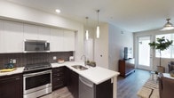 Located at 2510 West Ranch in Carlsbad, this model unit offers a thoughtfully designed single-floor layout that blends contemporary style with functional living. The open-concept floor plan allows the kitchen and living area to flow seamlessly into one another, creating a spacious and inviting atmosphere.

The kitchen is a standout feature, boasting modern dark cabinetry contrasted by crisp white upper cabinets and clean quartz countertops. A peninsula with an undermount stainless steel sink doubles as a breakfast bar, illuminated by elegant pendant lighting that adds warmth and visual interest. Stainless steel appliances, including a range, microwave, and dishwasher, complement the dark tile backsplash, lending a polished, upscale feel. Wood-look flooring extends throughout, adding warmth and continuity to the space.

The adjacent living area is open and bright, benefiting from recessed lighting and a neutral color palette that enhances the sense of airiness. A flat-screen television setup and warm-toned media console anchor the living space, creating a comfortable and stylish environment. The overall design reflects a cohesive modern aesthetic with high-quality materials and attention to detail throughout.