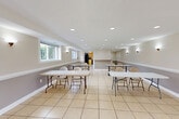 Located at 3308 Lockheed Blvd in Alexandria, Virginia, this single-floor community space offers a well-organized layout designed for gatherings and group activities. The interior is finished with large-format light ceramic tile flooring throughout, creating a clean, cohesive aesthetic that extends across every room.

The expansive dining area serves as the heart of the space, accommodating multiple long folding tables and chairs arranged for group use. Recessed lighting illuminates the room evenly from above, while wall-mounted sconces add a warm ambient glow. A two-tone wall treatment — neutral beige above and gray below a chair rail — adds a subtle architectural detail. Multiple windows fitted with security grilles allow natural light to filter in, offering views of the surrounding greenery. A glass door at one end opens to an outdoor area, enhancing the connection to the exterior.

The kitchen is equipped with light wood cabinetry, black appliances including a refrigerator, gas range, dishwasher, and microwave, along with a countertop workspace and a drop-ceiling with panel lighting. A dedicated service counter with open shelving separates the kitchen zone from the dining area, supporting event and catering needs. A connecting hallway provides additional circulation throughout the space.