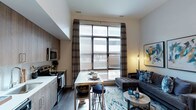 This contemporary single-floor residence showcases a thoughtfully designed open-concept layout that seamlessly blends the kitchen, dining, and living areas into one cohesive living space. Soaring ceilings amplify the sense of volume and airiness throughout, while oversized windows dressed with patterned curtains and horizontal blinds invite generous amounts of natural light. The kitchen is outfitted with modern stainless steel appliances, including a range and microwave, complemented by sleek flat-front cabinetry in a warm wood tone and a clean white countertop. A compact dining area with bar-height seating bridges the kitchen and living zone, making it ideal for casual meals or entertaining. The living area is anchored by a plush sofa and a round coffee table atop a vibrant patterned area rug, creating a welcoming and stylish retreat. Dark hardwood-style flooring runs throughout, providing a rich contrast to the light-toned walls and adding warmth to the overall aesthetic. The combination of high ceilings, expansive windows, and an efficient open floor plan gives this residence a spacious and modern feel that is both functional and visually striking.