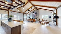 Located at 1021 Solace Pond View in Colorado Springs, CO, this single-floor space is a masterclass in modern rustic design. The open-concept layout seamlessly connects the kitchen, living, and dining areas beneath dramatically vaulted ceilings lined with natural wood planking and anchored by bold, dark-stained exposed timber beams. Travertine tile flooring flows throughout, accented by a decorative mosaic tile border that defines each functional zone.

The kitchen is a standout feature, boasting a generous white quartz island with bar seating, rich dark wood cabinetry, stainless steel appliances, and an artistic cluster of globe and cylinder pendant lights that cascade dramatically from the ceiling above.

The living area centers around a floor-to-ceiling whitewashed stone fireplace, which serves as a striking focal point beneath the soaring roofline. Curated open shelving, a wall-mounted display area, and thoughtfully placed seating create a welcoming and functional atmosphere. A dedicated dining nook with a round table and modern chairs sits beneath a vaulted alcove, bathed in natural light from multiple windows offering views of the surrounding Colorado landscape.

A connecting hallway leads further into the space, maintaining the clean, airy aesthetic found throughout. The overall ambiance is warm, open, and contemporary.