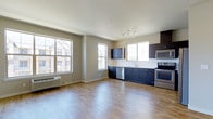 This single-floor space offers a thoughtfully designed open-concept layout that seamlessly integrates the living and kitchen areas into one cohesive environment. Large windows along the exterior wall flood the space with abundant natural light, creating an airy and welcoming atmosphere while offering views of the surrounding neighborhood.

The kitchen is a standout feature, outfitted with dark navy cabinetry that provides a striking contrast against the white countertops and neutral-toned walls. Stainless steel appliances — including a range, over-the-range microwave, and dishwasher — lend a modern, functional appeal. A track lighting fixture above the kitchen area provides focused illumination to complement the natural light.

The open living area extends generously from the kitchen, finished with warm hardwood-style flooring that runs throughout the entire space, adding continuity and a clean aesthetic. Neutral beige walls and white ceilings maintain a bright, versatile backdrop suitable for a variety of décor styles. A wall-mounted HVAC unit is conveniently positioned near the windows, ensuring year-round comfort. The overall design is contemporary and practical, making efficient use of the available space while maintaining a clean, modern look.