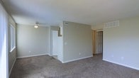 This single-floor space features an open main living area finished with wall-to-wall carpet and neutral beige-toned walls, creating a clean and understated atmosphere throughout. The living area is equipped with a ceiling fan and light fixture, providing both air circulation and overhead illumination. A textured ceiling adds subtle dimension to the space.

A central partial wall divides the main living area, defining separate zones while maintaining an open feel. HVAC vents are positioned throughout, including on the walls and ceiling, ensuring consistent climate control across the space. White baseboards run along the perimeter of the rooms, providing a crisp contrast to the neutral walls and carpeted floors.

Multiple doorways are visible from the main area, indicating access to additional rooms. One door leads to what appears to be a closet or utility space, while another open doorway leads to a connecting room or hallway, suggesting a functional multi-room layout. The overall design is practical and straightforward, with a simple, neutral palette that provides a versatile backdrop suitable for a variety of furnishing styles.