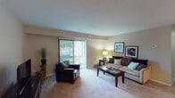 This single-floor residence offers a thoughtfully designed layout with distinct living, dining, kitchen, bedroom, and bathroom spaces, all connected by a functional flow visible in the overhead dollhouse view. The living room is a comfortable, carpeted space anchored by a sliding glass door fitted with vertical blinds, allowing natural light to filter in while providing access to an outdoor balcony with greenery views. A sofa, armchair, and glass-top coffee table furnish the area warmly. Adjacent to the living room, the dining area features hardwood-style flooring, a ceiling fan with integrated lighting, and space for a full dining set. The galley-style kitchen is outfitted with dark espresso cabinetry, granite-look countertops, a double basin sink, and black appliances, including a gas range and refrigerator. Two bedrooms offer carpeted floors and closet storage; the primary bedroom connects directly to an en-suite bathroom and features a spacious layout. Both bathrooms are finished with dark wood vanities, granite-look countertops, and large framed mirrors. A dedicated in-unit laundry area with a full-size washer and dryer adds convenience.
