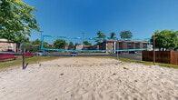 This outdoor recreational area features a full-size sand volleyball court as its centerpiece, offering residents a fun and engaging amenity in a comfortable community setting. The court is laid out on a generous expanse of fine sand, framed by a regulation-height net branded with colorful markings, and bordered by boundary lines. Surrounding the court, mature deciduous trees provide natural shade and a sense of seclusion, while well-maintained grassy lawn areas create a pleasant transition between the court and the adjacent multi-story brick residential buildings. Red benches are positioned along the edges of the sand area, offering convenient seating for spectators or those waiting to play. The community grounds feature paved parking areas nearby, along with additional landscaping that contributes to the overall green and inviting atmosphere. The buildings visible from the court display classic brick and panel exterior construction with private balconies, adding an architectural backdrop to the recreational space. Under open skies and surrounded by abundant greenery, this sand volleyball court serves as a vibrant outdoor gathering spot within the residential community, ideal for active recreation and social interaction among residents.