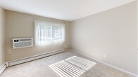 Located at 4 Silver Dr in Nashua, NH, this one-bedroom, one-bathroom unit offers a thoughtfully arranged single-floor layout. The bedroom is generously sized, featuring soft wall-to-wall carpeting, neutral beige walls, and a large window dressed with vertical blinds that allows natural light to pour in and cast bright patterns across the floor. A wall-mounted air conditioning unit and baseboard heating provide year-round climate control. The living area flows seamlessly from the bedroom, with the same neutral carpeting extending throughout, and features a distinctive pass-through window that opens to the kitchen, enhancing the sense of openness. The kitchen is equipped with white cabinetry, a dark refrigerator, and a compact countertop workspace, all visible through the pass-through opening. A hallway connects the main living spaces to the bathroom and entry doors, offering convenient access throughout the unit. The bathroom is clean and modern, showcasing white shaker-style cabinetry, a white countertop vanity, a framed mirror with overhead lighting, and wood-look flooring that adds warmth. A shower-tub combination completes the bathroom. Overall, the unit presents a functional, well-maintained space with a neutral palette suited for comfortable everyday living.