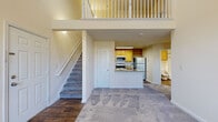 Located at 22541 Hickory Hill Square in Ashburn, Virginia, this thoughtfully designed residential space offers an open and airy floor plan highlighted by dramatic vaulted ceilings in the main living area. The living room is generously sized, featuring plush carpet, neutral walls, and large windows alongside a glass door that flood the space with natural light and provide outdoor views — all enhanced by a ceiling fan overhead.

The kitchen is well-appointed with warm honey-toned cabinetry, granite countertops, stainless steel appliances including a gas range, microwave, and refrigerator, as well as a peninsula with a sink that opens toward the living area. Rich wood-look flooring runs throughout the kitchen, adding warmth and durability.

A carpeted staircase leads to an upper loft landing with a white balustrade railing that overlooks the main living space below, adding architectural interest and a sense of vertical dimension.

The bedroom features soft carpeting, a ceiling fan, and a window with outdoor views, while a spacious walk-in closet provides ample storage. The bathroom is clean and functional, complete with a wood-vanity, tub-shower combination with tiled surround, and wood-look flooring. This home offers a comfortable, well-organized layout suited for everyday living.