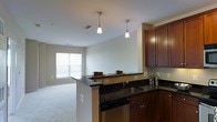 This single-floor residence offers a thoughtfully designed open-concept layout that seamlessly connects the kitchen and living area, creating a bright and welcoming atmosphere. The kitchen is a standout feature, showcasing rich dark wood cabinetry with sleek hardware, deep black granite countertops, and a peninsula that doubles as a breakfast bar — ideal for casual dining or entertaining. A stainless steel sink and dishwasher add a contemporary, functional edge to the space. Overhead, two elegant pendant lights hang above the peninsula, providing focused task lighting while contributing to the room's modern aesthetic. The open flow leads naturally into the living area, which is bathed in natural light streaming through a large window fitted with horizontal blinds. Neutral-toned walls and light carpeting in the living space create a calm, versatile backdrop that complements the kitchen's darker finishes. The overall ambiance strikes a balance between modern sophistication and comfortable everyday living, making it a highly functional and visually cohesive space throughout.