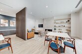 This single-floor communal space spans a generously sized open-plan layout integrating a living room, dining area, and fully equipped kitchen. The interior design follows a contemporary mid-century modern aesthetic, characterized by light hardwood flooring—laid in a striking chevron pattern in the kitchen and dining zone—white walls, and exposed wood beam accents overhead.

A slatted wood partition elegantly separates the living area from the dining space, maintaining visual connectivity while defining distinct zones. The living room features a marble-surround fireplace flanked by a wall-mounted TV, complemented by a patterned area rug, an orange leather sofa, and mid-century accent chairs. Built-in open shelving provides display and storage space along one wall.

The kitchen showcases dark wood cabinetry with brushed gold hardware, expansive quartz countertops, a large island with bar seating, and a marble tile backsplash. Pendant lighting with gold-toned fixtures adds warmth overhead.

Floor-to-ceiling black-framed sliding glass doors in the dining area flood the space with natural light and open directly to an outdoor courtyard and pool area, creating a seamless indoor-outdoor connection throughout.