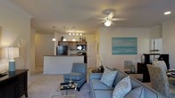 This single-floor space offers an open-concept layout that seamlessly connects the living area and kitchen. The living room is furnished with a plush sofa and accent chair in soft blue-grey tones, complemented by animal-print throw pillows that add a touch of personality. A glass-top coffee table sits atop a light area rug, grounding the seating arrangement. A ceiling fan with an integrated light fixture provides both ventilation and ambient lighting overhead. A large teal-toned wall canvas adds a serene, coastal aesthetic to the space.

The kitchen is visible from the living area, separated by a raised breakfast bar that doubles as a casual dining counter. Dark wood cabinetry provides ample storage, while black appliances, including a refrigerator and range, offer a functional contrast. A track lighting fixture and pendant lights illuminate the kitchen workspace effectively.

Neutral-toned carpet in the living area and light-colored walls throughout contribute to a bright, airy ambiance. A closet door and a window unit air conditioner are also visible, adding to the practical features of this well-appointed, comfortable space.