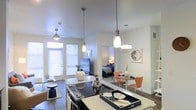 This single-story residence features an open-concept layout that integrates the kitchen, dining, and living areas into one cohesive, light-filled space. The kitchen is equipped with a sleek black electric cooktop set into a light-colored countertop, accented by stylish pendant lights that hang at varying heights, adding visual interest overhead. A ceiling fan in the living area contributes to the airy atmosphere. Large windows dressed with horizontal blinds line the far wall of the living area, allowing abundant natural light to fill the space while framing views of greenery outside. Dark hardwood-style flooring runs throughout, grounding the bright white walls and creating a clean contrast. A compact dining area sits adjacent to the kitchen, furnished with a round table and warm-toned orange chairs, adding a pop of color. A doorway from the main living space leads directly into the bedroom, which is softly lit and features a neatly arranged bed, maintaining the overall sense of openness and connectivity between rooms. The overall ambiance is modern, clean, and welcoming.