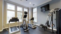 Located at 10 Primrose in Haverhill, Massachusetts, this fitness space offers a well-designed environment for exercise and wellness. The room is finished with a cool blue-gray color palette on the walls, complemented by crisp white crown molding and trim that lends the space a clean, polished aesthetic. Large windows with white-trimmed frames line the walls, allowing abundant natural light to fill the room while providing views of the surrounding outdoor greenery. The flooring consists of durable dark rubber or foam interlocking tiles, ideal for supporting heavy fitness equipment and providing comfort during workouts. The space is equipped with professional-grade Matrix brand cardio machines, including treadmills, an upright stationary bike, and an elliptical or stair-stepper, alongside a strength and rowing machine. A wall-mounted flat-screen television is positioned for easy viewing during workouts. Recessed lighting and a ceiling fan with a light fixture ensure the space remains comfortable and well-lit regardless of the time of day. The combination of ample natural light, quality equipment, and refined architectural details creates a motivating and functional fitness environment.