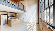 Located at 5000 Euclid Avenue in Cleveland, Ohio, this two-story industrial loft masterfully blends raw architectural character with modern finishes. The main level opens into an expansive living and kitchen area with polished concrete floors, soaring double-height ceilings, and dramatic floor-to-ceiling factory-style windows that flood the space with natural light. Exposed brick walls and a bold mezzanine level overhead add to the loft's industrial identity.

The kitchen is thoughtfully designed with warm wood cabinetry, stainless steel appliances including a refrigerator, range, and microwave, a sleek white quartz peninsula with an integrated sink, and pendant lighting overhead. Track lighting and dark accents throughout provide a refined contrast to the natural materials.

A striking black steel staircase leads to the upper level, where warm carpeting, exposed wooden beam ceilings, and industrial ductwork create an intimate yet characterful atmosphere. The bedroom is generously sized and features ample closet space. Both bathrooms are cleanly finished with white tile, wood-toned vanities, illuminated mirrors, and matte black fixtures. A dedicated laundry area and utility room add practical convenience to this architecturally distinctive urban loft.