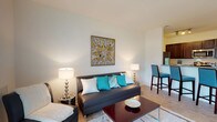 This well-designed single-floor residence offers a thoughtfully arranged layout that balances comfort and functionality across its living spaces. The entryway, finished with light wood-look flooring, opens to a clean, neutral palette that carries throughout the home.

The living area is bright and inviting, featuring light wood-look flooring, white walls, and large sliding glass doors that flood the space with natural light while providing views of the exterior. A designated dining area with a glass-top table and pendant lighting sits adjacent to the living room, creating a seamless open-plan flow.

The kitchen is equipped with dark espresso cabinetry, granite-style countertops, stainless steel appliances including a refrigerator and dishwasher, and light wood-look flooring that complements the rest of the home. A breakfast bar with bar seating connects the kitchen to the main living area.

The bedroom offers plush carpeting, a window with natural light, and ample space for furnishings. It is complemented by a full bathroom featuring large-format tile flooring, a dark vanity with a white countertop, a bathtub, and clean white walls. Throughout the residence, the neutral tones and consistent finishes contribute to a cohesive, contemporary ambiance.