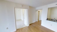 This single-floor residential space offers a clean, minimal aesthetic defined by its bright white walls and warm, light-toned hardwood flooring that runs through the main living areas. The layout is open and practical, with a hallway connecting the key areas of the home. A carpeted bedroom is accessible through a doorway off the main corridor, providing a softer, more comfortable underfoot feel in contrast to the hard flooring elsewhere. A paneled closet door lines the hallway, offering convenient built-in storage. The textured ceiling adds subtle dimension to the otherwise smooth, neutral interior. Natural light filters through the space, enhancing its airy and welcoming atmosphere. The consistent white trim and baseboards throughout contribute to a cohesive, well-maintained appearance. The overall ambiance is calm and versatile, offering a blank canvas that adapts easily to a variety of interior styles and furnishing preferences.