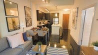 This single-floor space showcases a thoughtfully designed open-concept layout where the living, dining, and kitchen areas flow naturally into one another, creating a cohesive and functional environment. The polished concrete floors run throughout, lending an industrial-modern aesthetic that anchors the entire space with a sleek, reflective finish.

The kitchen is a standout feature, outfitted with rich dark cabinetry that reaches toward the ceiling and provides generous storage. Stainless steel appliances, including a full-size refrigerator and microwave, offer modern utility. A central island with a light-toned countertop and bar seating serves as both a prep area and a casual gathering spot. Pendant lighting above the island adds warm, focused illumination, complementing the track lighting overhead.

Adjacent to the kitchen, the dining area comfortably accommodates a full table and chairs, making it ideal for everyday meals or entertaining. The living area is defined by a striped area rug, a comfortable sofa, and a coffee table, creating an inviting lounge space. Wall art and warm ambient lighting contribute to the overall modern yet welcoming atmosphere of the space.