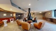 Located at 12520 Camino Del Sur in San Diego's Rancho Bernardo area, this thoughtfully designed communal lounge offers a generously sized, single-floor living space that blends contemporary aesthetics with inviting comfort. At the heart of the room stands a dramatic suspended black cone fireplace, serving as a bold focal point around which a curated seating arrangement is organized — including a deep charcoal sofa, curved cognac leather chairs, and a round low-profile coffee table. Polished terrazzo flooring runs throughout, lending a clean, upscale texture to the space. A series of tall arched gold-framed mirrors with hanging planters act as an elegant room divider, separating the lounge from a dining area furnished with a marble-top table and emerald green chairs. Along one wall, built-in cabinetry with warm wood tones provides storage beneath artful wall-mounted lighting and a curated art piece. Floor-to-ceiling glass doors flood the space with natural light and open directly to an outdoor courtyard, while hanging wicker swing chairs add a playful, resort-like touch near the entrance. A built-in wet bar and beverage station near the exterior doors adds functional convenience, making this lounge ideal for gathering and socializing.