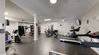 This fitness center offers a generously proportioned, single-floor workout space designed to accommodate a comprehensive range of exercise activities. The room features dark rubber flooring throughout, providing a durable and slip-resistant surface ideal for high-traffic gym use. White-painted walls and crown molding lend a polished, refined aesthetic, while decorative white columns add architectural character without interrupting the open layout.

The facility is equipped with an extensive selection of cardio machines, including multiple treadmills and elliptical trainers, as well as recumbent and upright stationary bikes. Strength training options include a multi-station cable machine, a power tower for bodyweight exercises, adjustable benches, and a well-stocked medicine ball rack featuring a colorful assortment of weighted balls. A large mirror on one wall visually expands the space and aids in form monitoring during workouts.

A wall-mounted flat-screen television provides entertainment during cardio sessions, and an inspirational wall decal reading "you are strong" adds a motivating touch. A water fountain is conveniently located within the space. Ceiling-mounted light fixtures and flush fluorescent lighting ensure the room is well-lit throughout. A windowed entry door allows a modest amount of natural light into the space.