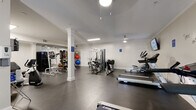 This fitness center offers a generously proportioned, single-floor workout space designed to accommodate a comprehensive range of exercise activities. The room features dark rubber flooring throughout, providing a durable and slip-resistant surface ideal for high-traffic gym use. White-painted walls and crown molding lend a polished, refined aesthetic, while decorative white columns add architectural character without interrupting the open layout.

The facility is equipped with an extensive selection of cardio machines, including multiple treadmills and elliptical trainers, as well as recumbent and upright stationary bikes. Strength training options include a multi-station cable machine, a power tower for bodyweight exercises, adjustable benches, and a well-stocked medicine ball rack featuring a colorful assortment of weighted balls. A large mirror on one wall visually expands the space and aids in form monitoring during workouts.

A wall-mounted flat-screen television provides entertainment during cardio sessions, and an inspirational wall decal reading "you are strong" adds a motivating touch. A water fountain is conveniently located within the space. Ceiling-mounted light fixtures and flush fluorescent lighting ensure the room is well-lit throughout. A windowed entry door allows a modest amount of natural light into the space.