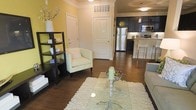 This single-floor residence offers an inviting open-concept layout that seamlessly connects the living, dining, and kitchen areas. The living space is anchored by rich, dark hardwood flooring and features warm yellow-toned walls accented by crisp white crown molding and trim, creating a cheerful yet polished ambiance. A plush white area rug, glass coffee table, and a light-colored armchair lend the space a clean, comfortable feel, while a dark open shelving unit provides both storage and display opportunities.

The kitchen is a standout feature, designed with dark cabinetry, stainless steel appliances including a refrigerator, microwave, and range, and a breakfast bar with modern bar stools. Pendant lighting above the bar area adds a touch of elegance to the functional space. The kitchen flows naturally into the living area, making it ideal for entertaining.

Two interior doors are visible from the main living area, suggesting additional private rooms beyond. The space benefits from recessed and flush-mount ceiling lighting throughout, ensuring a well-lit environment at any time of day. Overall, the residence combines modern finishes with warm, welcoming design elements to create a cohesive and practical living environment.