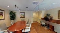 Located at 6021 Crape Myrtle Ct in Los Angeles, CA, this single-floor professional office space is thoughtfully designed to accommodate both collaborative and individual work needs. The layout is divided into two primary functional zones, separated by a full-height glass partition wall that maintains an open, transparent feel throughout the space.

The conference area is anchored by a substantial oval wood conference table surrounded by cushioned cream-colored chairs, providing a polished setting for meetings. Track lighting mounted on the ceiling supplements recessed can lights, ensuring the room is well-illuminated. Decorative framed landscape artwork and lush indoor plants add warmth to the otherwise professional environment. Patterned carpet in warm earth tones runs throughout the space, contributing to its inviting atmosphere.

Adjacent to the conference zone, a dedicated workstation area features a built-in cherry wood desk running along the wall, equipped with multiple computer terminals and ergonomic seating. Framed artwork and additional greenery maintain visual continuity across both areas.

Large floor-to-ceiling windows and glass entry doors at the front of the space flood the interior with natural light and offer clear views of the landscaped exterior. The overall ambiance is professional yet approachable, combining functional office design with tasteful decorative elements.