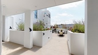 This rooftop terrace is a thoughtfully designed outdoor amenity space that combines artistic expression with functional outdoor living. The terrace is divided into several distinct zones, each offering a unique experience while maintaining a cohesive modern aesthetic defined by clean white walls, concrete flooring, and open sky views.

A covered entry area with white structural columns leads into the main terrace, where decorative bird silhouettes in black and vibrant colors are mounted across the white exterior walls, creating a striking artistic focal point. String lights are strung overhead, adding a festive ambiance suited for evening gatherings. Raised planters throughout the space are filled with flowering plants, succulents, and trailing greenery, softening the contemporary architecture with natural elements.

One section of the terrace features a pergola with a slatted white roof structure, providing partial shade over an outdoor kitchen equipped with a built-in grill and bold patterned tile detailing along a built-in bench. An adjacent lounge area under the same pergola centers around a sleek fire pit table, flanked by generous cushioned seating with striped upholstery. Additional dining tables and chairs are positioned throughout, accommodating both intimate and larger gatherings beneath the open sky.