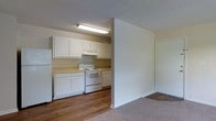 This single-story space features an open-concept layout that connects the kitchen and living area through a half-wall partition, creating a functional yet defined separation between the two zones. The kitchen is equipped with white raised-panel cabinetry spanning both upper and lower sections, providing ample storage. A white electric range and a full-sized refrigerator are included, complemented by light-toned countertops. A fluorescent ceiling fixture mounted beneath the upper cabinets ensures bright, even task lighting throughout the kitchen. The flooring in the kitchen area consists of wood-look planks, adding a warm, durable surface suited to high-use areas. The adjacent living space transitions to carpeted flooring, offering a softer underfoot feel. White walls and white trim throughout contribute to a clean, cohesive aesthetic. A textured popcorn ceiling is present in both areas. A door visible along the far wall leads to an additional room or closet. Overall, the space maintains a straightforward, functional design with neutral finishes that provide a versatile backdrop for furnishing and personalization.