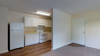 This single-story space features an open-concept layout that connects the kitchen and living area through a half-wall partition, creating a functional yet defined separation between the two zones. The kitchen is equipped with white raised-panel cabinetry spanning both upper and lower sections, providing ample storage. A white electric range and a full-sized refrigerator are included, complemented by light-toned countertops. A fluorescent ceiling fixture mounted beneath the upper cabinets ensures bright, even task lighting throughout the kitchen. The flooring in the kitchen area consists of wood-look planks, adding a warm, durable surface suited to high-use areas. The adjacent living space transitions to carpeted flooring, offering a softer underfoot feel. White walls and white trim throughout contribute to a clean, cohesive aesthetic. A textured popcorn ceiling is present in both areas. A door visible along the far wall leads to an additional room or closet. Overall, the space maintains a straightforward, functional design with neutral finishes that provide a versatile backdrop for furnishing and personalization.