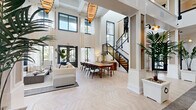 Located at 5201 W Hillsboro Blvd in Coconut Creek, Florida, this stunning single-floor clubhouse spans a generous footprint across five distinct areas, delivering an upscale, resort-inspired experience. The grand entry foyer sets an immediate tone of elegance, showcasing soaring double-height ceilings, dramatic floor-to-ceiling windows that flood the space with natural light, and a striking herringbone-patterned light wood floor that flows throughout. An ornate area rug anchors the entry, complemented by lush tropical potted palms and a statement wood-paneled sliding door framed in polished brass hardware. The central great room opens expansively, featuring a sleek black staircase with metal railings, woven pendant light fixtures, a fireplace with a bold botanical mural accent wall, and a built-in window nook. A dedicated conference and media room offers a sophisticated atmosphere with coffered ceilings, ambient LED lighting, bold tree-patterned wallcovering, a large-format wall-mounted display, and a private adjacent office. A vibrant lounge area showcases teal velvet banquette seating, eclectic colorful pendant lights, tropical mural wallpaper, and leather accent chairs, creating a lively yet refined social retreat. Throughout, the design masterfully balances contemporary elegance with warm, tropical Florida-inspired character.