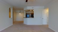 This single-floor residence offers a well-designed layout that balances openness with defined living spaces. The main living area is generously proportioned, featuring neutral carpet flooring, clean white walls, and a ceiling fan that adds both function and a classic aesthetic touch. A graceful arched doorway connects the living space to a windowed alcove, where a large multi-pane window fills the area with natural light, creating a bright and welcoming atmosphere.

The kitchen is thoughtfully designed with dark wood cabinetry, built-in black appliances, and a wraparound breakfast bar that seamlessly integrates the kitchen with the adjacent living and dining areas. Crown molding detail frames the kitchen's upper structure, adding an elegant architectural accent. A pendant light fixture hangs above, casting warm ambient light over the space. The open-concept flow between the kitchen, dining, and living areas makes the layout ideal for everyday living and entertaining alike.

Throughout the home, consistent neutral tones and soft carpeting contribute to a calm, cohesive ambiance. Louvered closet doors and recessed vents reflect practical, well-considered design details. Overall, this space combines comfort, functionality, and understated style across its single-floor footprint.