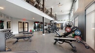 Located at Park Meadows Drive in Lone Tree, Colorado, this two-story fitness facility delivers a comprehensive workout environment spread across two generously sized floors. The main level commands attention with its soaring double-height ceilings, abundant natural light streaming through oversized windows, and durable speckled rubber flooring throughout. This floor is dedicated to strength training and functional fitness, housing Rogue power racks, cable machines, a full dumbbell rack, weight benches, and a variety of selectorized resistance machines. A dedicated room off the main floor offers TRX suspension trainers, rowing machines, and floor-to-ceiling mirrors — ideal for functional training and recovery work. An elegant staircase with dark metal railings connects the levels, and a mezzanine-style open railing allows views between floors. The upper level is home to a dedicated cardio suite featuring rows of treadmills and Echelon spin bikes, each equipped with personal screens. Elliptical trainers and stair climbers round out the upper level's offerings. Large windows line the upper floor, providing natural light and exterior views. The facility's modern aesthetic is defined by a neutral gray and black color palette, recessed and pendant lighting, and clean architectural lines throughout.
