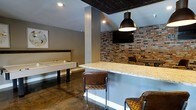 This single-floor entertainment space is thoughtfully designed to create an inviting and energetic atmosphere for socializing and recreation. The room immediately captures attention with its striking exposed brick accent wall, which adds warmth and an industrial character to the overall aesthetic. Complementing this is a dramatic dark-painted ceiling that lends depth and a modern edge to the space.

A central granite-topped bar counter serves as the focal point of the room, flanked by leather-upholstered bar stools that provide comfortable seating for guests. Industrial-style pendant lighting hangs above the bar, casting a warm glow across the counter and enhancing the lounge ambiance. A wall-mounted flat-screen television positioned above the brick wall ensures entertainment is always within view.

The game area features an air hockey table, offering a fun and interactive element for visitors. The polished concrete flooring throughout the space is both durable and visually cohesive, tying together the industrial and contemporary design elements. Recessed lighting supplements the pendant fixtures, ensuring the space is well-lit throughout. The neutral gray walls provide a versatile backdrop that allows the brick, dark ceiling, and warm furnishings to stand out, making this a well-balanced and engaging entertainment environment.