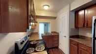 This two-story residence in Charlestown offers a well-organized layout across two floors. The kitchen is a functional centerpiece, showcasing rich medium-toned wood cabinetry that extends to the upper walls, providing generous storage. Dark granite-style countertops contrast warmly against the cabinetry, lending the space a classic, grounded aesthetic. The kitchen is equipped with an electric coil range and a double-basin stainless steel sink positioned beneath a window, allowing natural light to filter in. A louvered door adds a traditional touch while offering practical utility for a pantry or closet. Hardwood-style flooring runs throughout, contributing to a cohesive and warm visual flow. An overhead range hood and flush-mount ceiling light provide both functional ventilation and ambient illumination. The open sightline through the kitchen toward an adjoining room—which benefits from additional natural light through vertical blinds—creates a connected, open feel. The two-floor layout provides defined separation between living and private areas, making the space both versatile and comfortable for everyday living.