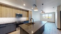 This contemporary single-floor space is designed with a clean, modern aesthetic that balances form and function throughout. The heart of the unit is a well-appointed open-concept kitchen, anchored by a generous island with a dark quartz countertop, an undermount sink, and a matte black faucet. Pendant lights hang above the island, providing both task lighting and a stylish focal point. The kitchen features warm natural wood upper cabinetry paired with slate-blue lower cabinets, a classic white subway tile backsplash, and a full suite of stainless steel appliances including a refrigerator, range, microwave, and dishwasher. Light hardwood flooring flows seamlessly throughout the space, adding warmth and continuity. The bedrooms are bright and airy, each benefiting from large windows that invite abundant natural light and offer views of the surrounding landscape, including distant mountains and greenery. Ceiling fans provide comfort in the sleeping areas. The bathroom is finished to a high standard, featuring a dark quartz vanity countertop, a large frameless mirror, matte black fixtures, and a white subway tile shower enclosure. Overall, the space presents a cohesive modern design with thoughtful finishes and a bright, open ambiance.