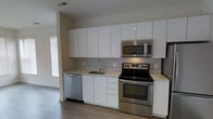 This single-floor space showcases a clean, modern aesthetic throughout. The kitchen is a standout feature, designed with a streamlined, galley-style layout that maximizes both functionality and visual appeal. Floor-to-ceiling white gloss cabinetry lines the walls, offering generous storage while contributing to a bright, airy atmosphere. The light-toned countertops provide ample prep space and complement the crisp white palette seamlessly. A full suite of stainless steel appliances — including an electric range, over-the-range microwave, and dishwasher — lends a polished, contemporary feel. The undermount sink is positioned beneath a modern faucet, adding a refined touch to the workspace. Hardwood-style flooring runs throughout, grounding the space with warmth and texture that contrasts elegantly with the white cabinetry. Natural light filters in through a nearby window, brightening the interior and enhancing the open, uncluttered ambiance. The visible vent and recessed ceiling details reflect thoughtful construction and practical design. Overall, this space presents a well-appointed, move-in-ready environment with a cohesive modern style suited to a variety of lifestyles.