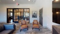 Located at 11000 Eagle Hall Ln in Goose Creek, South Carolina, this two-story amenity building showcases a refined modern farmhouse aesthetic throughout its thoughtfully designed interior. Wide-plank hardwood flooring, crisp shiplap-clad walls, and dark barn-style doors establish a cohesive design language that flows across both levels.

The ground floor opens to multiple distinct lounge areas, including a seating nook with shiplap walls, framed artwork, and a wall-mounted TV, as well as a more intimate space featuring rich leather armchairs and an oversized display screen paired with a rustic wood media console. Steel-framed glass partition walls allow natural light to move freely between spaces while maintaining visual separation. One sunlit room boasts floor-to-ceiling black-framed windows, exposed wood ceiling beams, and bold green lounge seating, creating a bright and inviting atmosphere with outdoor views.

A business center offers workstations with computers, a printer, and ample natural light from a generous window wall. The staircase connects both floors beneath a striking lantern-style chandelier, flanked by bold dark horizontal railings. The upper level continues the design motif, featuring decorative pressed-tin accent walls, additional seating areas, and open sightlines to the floor below, reinforcing the building's spacious, welcoming character.