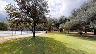 This expansive outdoor space presents a well-maintained, park-like environment characterized by a generous open lawn area shaded by several large, mature trees. The canopy of established deciduous and broadleaf trees provides natural shade across the grassy grounds, creating a comfortable and inviting atmosphere throughout the space. Fallen leaves scattered across the lawn suggest a seasonally dynamic environment that changes character throughout the year.

To one side, a fenced tennis court is visible, indicating that the space serves both recreational and leisure purposes. A gravel or paved pathway runs alongside the court, offering a defined route through the grounds. Benches are positioned along the path, providing spots to rest and enjoy the surroundings.

In the background, multi-story brick buildings frame the space, suggesting a campus or institutional setting. The architecture of these structures complements the outdoor environment with a traditional, established aesthetic. Additional amenities such as bike racks and waste bins are noted near the buildings, reinforcing the functional nature of the space. Under a partly cloudy sky, the grounds benefit from generous natural light, enhancing the open, airy ambiance of this versatile outdoor area.