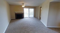 This single-floor space centers around a spacious living room that combines comfort and functionality. The room is anchored by a classic brick fireplace with a white-painted mantel, serving as a focal point along one wall. A ceiling fan with an integrated light fixture is mounted overhead, providing both air circulation and ambient lighting.

Natural light flows generously into the space through a large sliding glass door, which opens to an outdoor area and offers pleasant exterior views. The neutral-toned carpeting extends throughout the room, contributing to a warm and understated aesthetic. The walls are painted in a soft, warm beige tone that complements the overall color palette.

A hallway visible along the right side of the living room leads further into the home, suggesting additional rooms beyond the main living area. The layout appears open and straightforward, with clean lines and minimal architectural complexity. White baseboards accent the perimeter of the room, adding a subtle contrast to the carpeting. Overall, the space presents a practical and livable environment suited for everyday use.