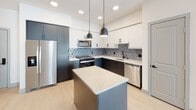 This thoughtfully designed single-floor residence showcases a clean, contemporary aesthetic throughout. The heart of the unit is an open-concept kitchen and living area, where light hardwood-style flooring flows seamlessly underfoot. The kitchen is a standout feature, outfitted with dark lower cabinetry contrasted by crisp white upper cabinets, a striking geometric-patterned tile backsplash, stainless steel appliances, and a center island with a light stone countertop. Sleek pendant lights with an industrial finish hang above the island, adding character to the space. The adjacent living area benefits from a large window that draws in natural light, and a ceiling fan overhead adds comfort.

The bedroom offers plush carpeting for a cozy feel, a ceiling fan, and two gray panel doors — one leading to a generous walk-in closet with built-in shelving and another to an additional space, providing ample storage.

The bathroom is finished with large-format tile flooring and a full tiled tub-and-shower surround in a soft gray linear pattern, complemented by matte black hardware accents and a curved shower rod. The overall palette across the unit is light and neutral, creating a bright, modern ambiance throughout.