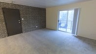 This single-floor space offers a straightforward yet characterful layout defined by a bold exposed brick accent wall that serves as the room's focal point. The dark, textured brickwork contrasts effectively with the smooth, light-toned walls on the adjacent sides, creating a visually dynamic interior. The flooring throughout is covered in neutral gray carpet, providing a soft and cohesive foundation that complements the mixed material palette of the room.

Natural light is introduced generously through a large sliding glass door positioned along the exterior wall. This door opens onto a balcony enclosed by a metal railing, where outdoor views of surrounding trees and landscape are visible, suggesting a serene setting. The sliding door allows for both ventilation and a seamless visual connection between the interior and the outdoors.

The ceiling is paneled in a light tone, contributing to a clean overhead finish, while the open floor plan provides flexibility in how the space can be arranged and utilized. Overall, the room presents a blend of rugged and refined elements, with the brick wall adding warmth and texture to an otherwise understated and versatile interior.