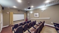 Located at 4020 Eight Belles Ln. in Greensboro, NC, this dedicated home theater room delivers an authentic cinema experience within a single-floor layout. The space is designed with tiered, stadium-style seating, featuring rows of plush brown fabric theater chairs with built-in cup holders and black armrests — accommodating a generous number of viewers in comfort. The seating is arranged on raised platforms, ensuring unobstructed sightlines from every seat toward the large, wall-mounted projection screen at the front of the room. A ceiling-mounted projector serves as the centerpiece of the audiovisual setup, complemented by surround-sound speakers strategically positioned throughout the space. The room is finished with warm taupe-toned walls adorned with framed black-and-white photography, lending a classic Hollywood ambiance. Recessed ceiling lighting and wall sconces provide soft, dimmable illumination suited for screening events. A charming red vintage-style popcorn machine stationed near the screen adds a playful, concession-stand touch. A built-in cabinet near the rear of the seating area provides storage and operational space. The flooring features a combination of deep red carpet on the tiered platforms and a patterned area rug at the front, completing the immersive, theater-inspired aesthetic.