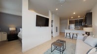 This single-floor unit showcases a contemporary, open-concept design with light hardwood-style flooring and neutral white walls throughout, creating a bright and cohesive atmosphere. Recessed lighting illuminates the main living and kitchen areas, while a ceiling fan adds comfort overhead.

The living area features a wall-mounted flat-screen television anchored to a structural column that subtly separates the sleeping space from the main living zone. A large window fills the seating area with natural light and offers outdoor views. Industrial-style nesting coffee tables and a soft area rug complete the lounge space.

The kitchen is well-appointed with dark espresso cabinetry, a textured tile backsplash, white countertops, and a full suite of stainless steel appliances, including a refrigerator, electric range, and microwave. A peninsula with bar stools provides casual dining and additional workspace.

Tucked conveniently between the living area and kitchen, a stacked washer and dryer unit offers in-unit laundry. The bathroom features a dark wood-grain vanity with a white countertop, chrome fixtures, and light flooring, maintaining the modern aesthetic carried throughout the space.