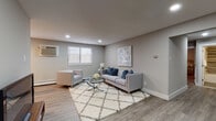 This thoughtfully designed single-floor residence offers a cohesive, contemporary aesthetic defined by neutral grey tones, clean white trim, and wood-look flooring that runs throughout. The open living area is generously proportioned, accommodating a full seating arrangement centered around a glass coffee table atop a diamond-patterned shag rug. Recessed lighting illuminates the space, while windows fitted with white blinds provide natural light. A wall-mounted air conditioning unit and baseboard heating ensure year-round comfort. A dedicated area for a television and media console anchors one side of the room, and a separate dining space is visible beyond.

The galley-style kitchen is efficiently laid out, featuring dark cabinetry, crisp white countertops, a stainless steel sink, and a navy blue tile backsplash. Stainless steel appliances, including a gas range, microwave, and refrigerator, complete the functional setup.

The primary bedroom offers a calm retreat with a ceiling fan, natural light from a window, and ample space for a full dresser and nightstands. The secondary bedroom is similarly finished with a ceiling fan, a closet with double doors, and a bright window. The bathroom features a dark vanity with a white countertop, mirrored medicine cabinet, and tile flooring.