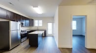 This single-story space at Windsor Castle showcases a thoughtfully designed interior with a strong emphasis on the kitchen as a central feature. The kitchen is outfitted with rich, dark espresso-toned cabinetry that extends to the upper and lower levels, providing generous storage. A central island with light granite-style countertops serves as both a prep space and a visual anchor for the room. Stainless steel appliances, including a gas range, microwave, and dishwasher, complement the contemporary aesthetic. The countertops along the perimeter mirror the island's stone finish, creating a cohesive look throughout. Wide-plank hardwood flooring in a warm medium tone runs continuously through the kitchen and into the adjoining areas, adding warmth and visual continuity. Natural light enters through multiple windows, including a glass-paneled exterior door, brightening the neutral cream-toned walls. The open layout allows for a smooth transition between the kitchen and the connected living areas, which are partially visible and share the same flooring. The overall ambiance is clean, modern, and functional.