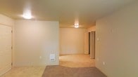 This single-floor space presents a straightforward, open layout with a clean and neutral aesthetic throughout. The main living area is finished with warm beige walls and standard-height ceilings, creating a modest yet comfortable atmosphere. A flush-mount ceiling light provides overhead illumination, supplementing the ambient lighting in the room.

The flooring transitions between tile and carpet, with the carpeted area defining the main living space and the tile visible near the entry or adjacent zone, offering a practical distinction between areas. A wall-mounted vent is positioned near the baseboard, indicating a built-in heating or ventilation system.

Toward the back of the space, a hallway leads to additional rooms, with what appears to be a door and a refrigerator visible, suggesting proximity to a kitchen or utility area. Multiple electrical outlets are distributed along the walls, offering convenience throughout the space.

Overall, the unit features a simple, functional design with neutral finishes that provide a blank canvas for personalization. The open feel of the main area and the accessible layout make it a practical and adaptable living space.