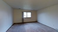 This single-floor space presents a straightforward and functional layout designed to offer a comfortable, livable environment. The main room is generously sized, featuring wall-to-wall carpeting in a neutral tone that adds warmth underfoot. The walls are finished in a light cream hue, creating a bright and airy atmosphere that feels open and welcoming. A textured ceiling adds subtle architectural character throughout the space. Natural light enters through a centrally positioned window fitted with vertical blinds, providing both illumination and privacy control. Recessed ceiling vents indicate a built-in HVAC system, ensuring year-round climate comfort. The room's clean, uncluttered design makes it highly versatile and easy to personalize. The neutral color palette throughout serves as an ideal canvas for a wide range of interior styles. The overall layout is practical and efficient, with smooth transitions between wall and floor surfaces defined by clean baseboards. This space is well-suited for those seeking a simple, move-in ready environment with a calm, understated aesthetic.