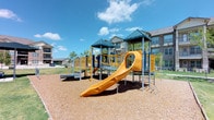 This outdoor playground offers a well-designed recreational space situated within a lush, manicured green lawn. The centerpiece of the space is a multi-tower play structure finished in bold yellow and navy blue, featuring a large curved slide, climbing ladders, bridged walkways, grid-panel railings, and a convex mirror for interactive play. The play equipment is set atop a generous wood chip safety surface, providing both cushioning and a defined play zone. Surrounding the structure, the open lawn extends across a spacious area with young trees planted throughout, offering growing shade and a sense of greenery. Concrete pathways wind through the grounds, providing clear and accessible routes for pedestrians. Ground-level bollard lighting is strategically placed along the walkways, supporting safe use during evening hours. Additional play panels and standalone activity elements are positioned around the perimeter of the main structure, adding variety for younger children. The playground is framed by multi-story residential buildings constructed with brick and stone facades, featuring covered balconies and pitched rooflines that contribute a cohesive architectural backdrop. The entire space enjoys open sky exposure, making it a bright and inviting environment for outdoor activity and community engagement.