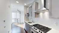 This contemporary single-floor residence is designed with clean lines and modern finishes throughout. The kitchen serves as a standout feature, showcasing two-tone cabinetry with soft gray lower cabinets and glass-front upper cabinets that add both style and functionality. A classic white subway tile backsplash runs the length of the cooking area, complementing the crisp white quartz countertops and stainless steel undermount sink. The kitchen is equipped with a professional-grade gas range, a built-in microwave, and a stainless steel range hood, making it a well-appointed space for cooking enthusiasts. Recessed lighting keeps the kitchen bright and inviting. Rich, dark hardwood flooring flows seamlessly from the kitchen into the adjacent living area, creating a cohesive and warm aesthetic. Floor-to-ceiling sliding glass doors in the living area flood the space with abundant natural light and provide direct access to an outdoor balcony, blurring the line between indoor and outdoor living. A ceiling fan enhances comfort in the main living space. The overall ambiance is modern and airy, with a neutral palette that offers a versatile canvas for any interior style.