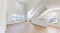 Located at 1 Avalon Drive in Hull, MA, this two-story home offers a well-designed layout across two floors. The main level immediately impresses with a dramatic double-height living area, where soaring ceilings and large windows — including a prominent upper-level window — flood the space with abundant natural light. Light-toned hardwood-style flooring runs throughout the main level, complementing the crisp white walls and creating a clean, airy aesthetic. A staircase with white balusters and a carpeted runner leads to the upper level, adding architectural interest to the open living area. The kitchen features white shaker-style cabinetry, stainless steel appliances including a refrigerator and dishwasher, and light countertops, offering both style and function. A dedicated laundry area with wire shelving provides added convenience. The bathroom is finished with a gray vanity, white quartz countertop, a single sink, and a large framed mirror. Upstairs, the generously sized primary bedroom offers ample space, while the second bedroom features light flooring and a window with views of the surrounding greenery. Two closets provide practical storage throughout. The home's neutral palette and consistent finishes create a cohesive, move-in ready environment.
